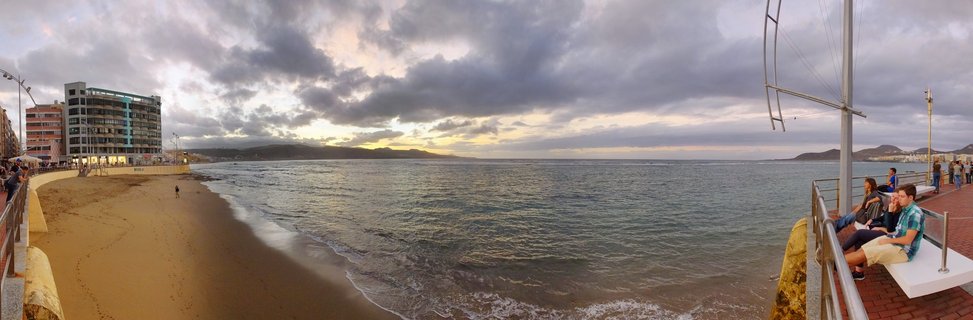 Wide coastal view of Las Canteras Beach showing sandy shoreline, ocean waves, dramatic cloudy sky at sunset, beachfront buildings, and people sitting on promenade benches