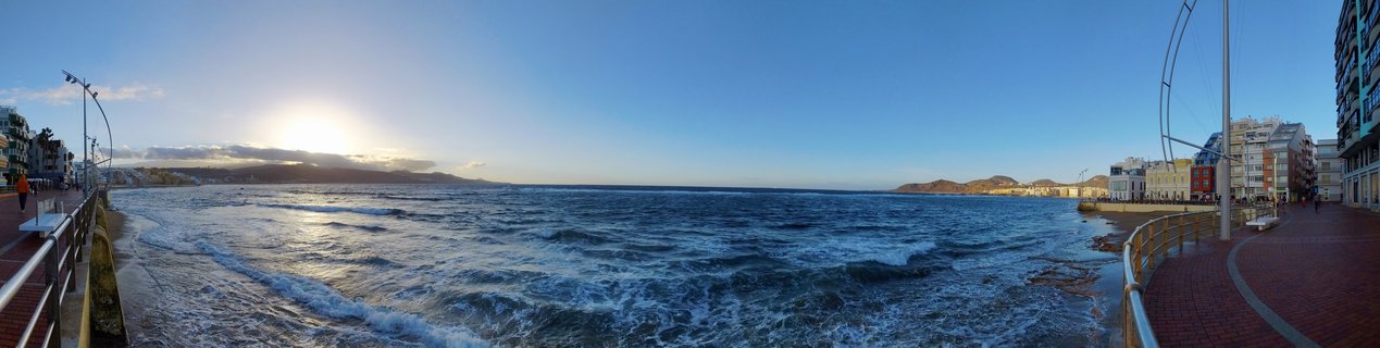 Wide panoramic photograph of Las Canteras beach showing the Atlantic Ocean with waves, curved promenade walkways on both sides, coastal buildings, distant mountains, and clear blue sky with low sun