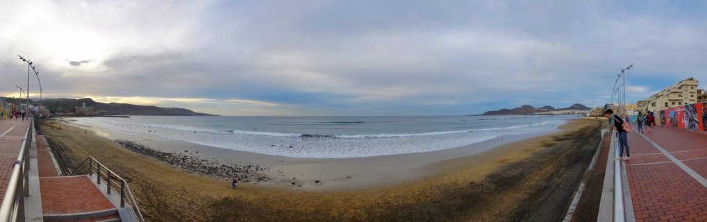 Wide panoramic view of Las Canteras Beach showing sandy shoreline, ocean waves, brick promenade with pedestrians, street lamps, buildings, and distant mountains under cloudy sky