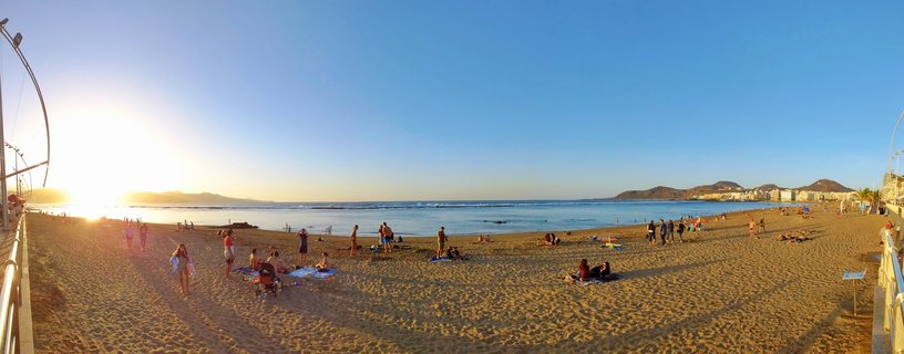 Panoramic view of Las Canteras Beach at sunset showing people relaxing on golden sand with ocean waves, distant mountains, and clear blue sky during golden hour in Gran Canaria, Spain