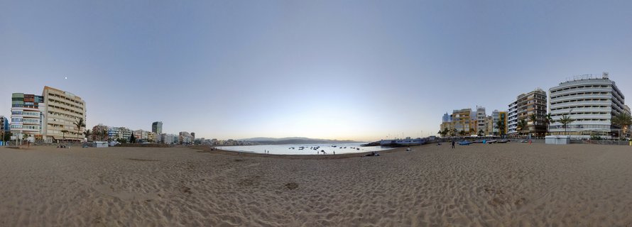 Wide panoramic view of Las Canteras beach showing golden sand foreground, calm bay with small boats, hotels and buildings along both sides of the coastline, clear sky at dusk