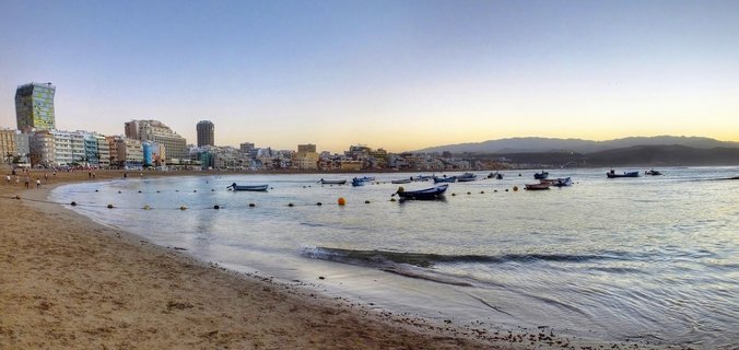 Panoramic view of Las Canteras Beach showing golden sand, calm blue water with small fishing boats and yellow buoys, hotels and buildings along the coastline, and mountains in the distance under a clear sunset sky