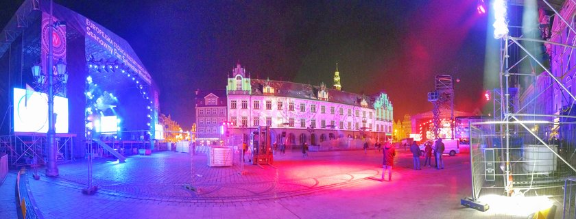Night photograph of Wroclaw Market Square showing a large event stage with blue lighting, historic buildings illuminated in pink and green, and people walking on cobblestone pavement