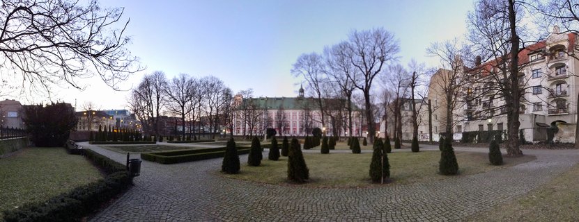 Panoramic view of formal gardens with cobblestone pathways and conical evergreen trees leading to the Royal Castle Poznań, a pink baroque palace with green roof, at winter twilight with bare trees and residential buildings visible, Poznań, Poland