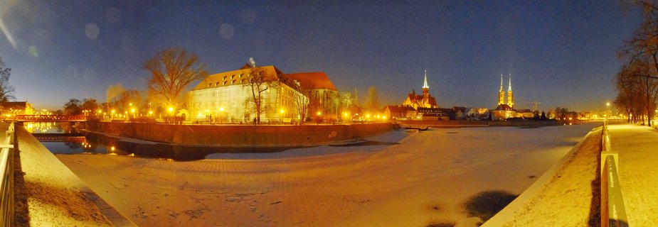 Panoramic night view of Wrocław, Poland showing a frozen river with illuminated historic buildings, cathedral spires, bare trees, and golden streetlights reflecting on ice and snow during winter twilight