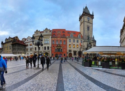 Wide view of Prague's Old Town Square showing the Gothic clock tower, colorful historic buildings with red roofs, cobblestone pavement, and pedestrians walking past market stalls under an overcast winter sky