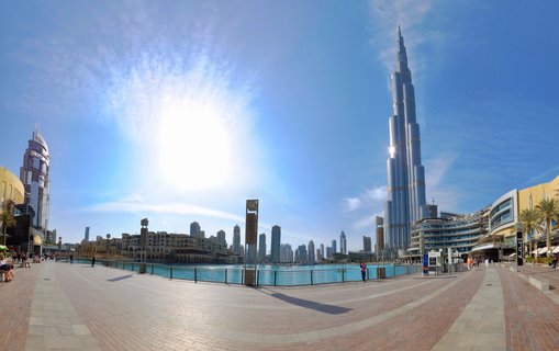 Panoramic view of Burj Khalifa skyscraper towering over Dubai Fountain with people walking on brick promenade under bright blue sky in Downtown Dubai, UAE