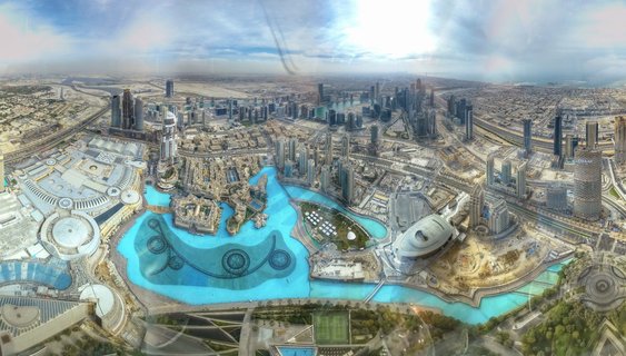 Aerial panoramic view of Downtown Dubai showing the Dubai Fountain's blue waters surrounded by skyscrapers, highways, and desert landscape extending to the horizon
