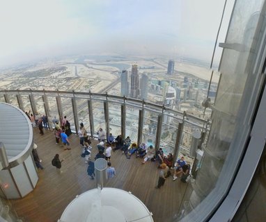 High-angle view of tourists standing and sitting on wooden observation deck inside Burj Khalifa, with Dubai cityscape, water canal, and desert visible through glass windows