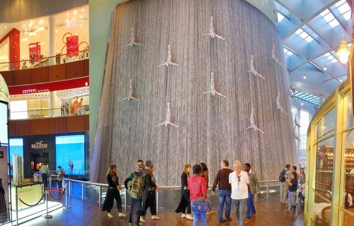 Indoor waterfall sculpture with bronze diving human figures at Dubai Mall, surrounded by visitors observing from ground level with shopping mall stores visible on multiple floors