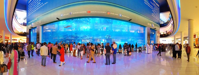 Large cylindrical aquarium tank in shopping mall with sharks and fish swimming in blue water, surrounded by diverse crowd of visitors watching from polished white marble floor