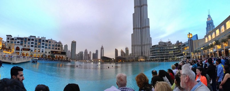 Wide panoramic view of Dubai Fountain lake with crowds of people in foreground, Burj Khalifa skyscraper rising prominently in center background, surrounding buildings and palm trees along waterfront at dusk