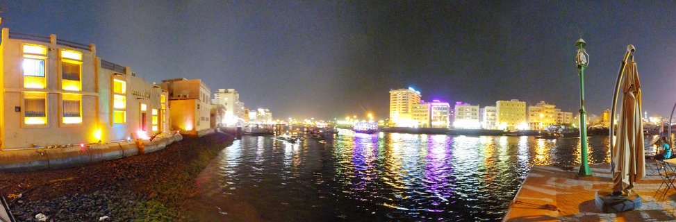 Night panoramic view of Dubai Creek showing traditional buildings with yellow lighting on the left, water with colorful reflections in the center, and modern illuminated buildings in the background with a waterfront promenade on the right
