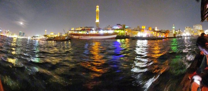Night panorama of Dubai Creek showing traditional wooden boats docked along the waterfront with an illuminated minaret tower and colorful building lights reflecting on dark water