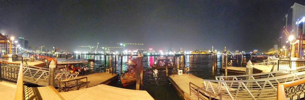 Night view of traditional wooden abra boats at Dubai Creek dock with passengers, illuminated buildings across the water, and city lights reflecting on the water surface