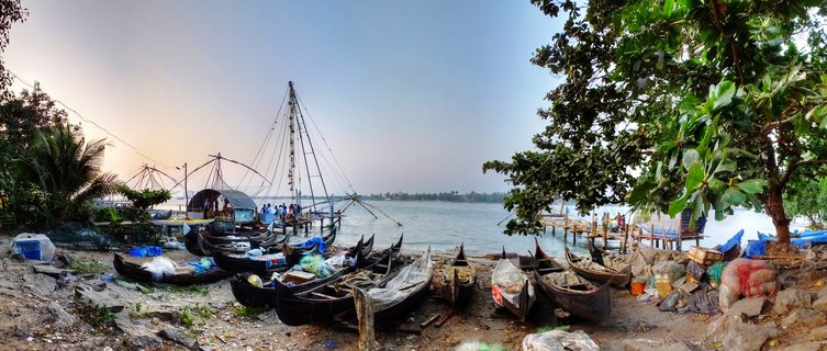 Multiple Chinese fishing nets and traditional wooden fishing boats on the shore at Fort Kochi harbor with palm trees and water in the background during golden hour