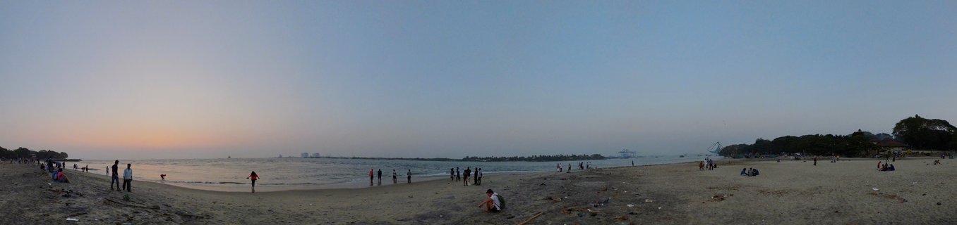 Wide panoramic view of a beach at dusk with numerous people walking and sitting on sand, ocean waves in the middle ground, and gradient sky from orange to blue above the horizon