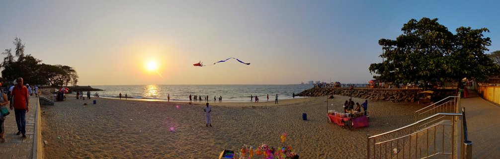 Panoramic beach scene at sunset showing people on sand, ocean with ships on horizon, colorful kite flying in sky, trees and vendor stalls along the shoreline in Kochi, India