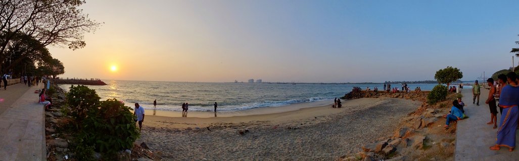 Panoramic view of a beach at sunset with people walking on sand and paved pathways, the sun setting over ocean water with distant buildings visible on the horizon