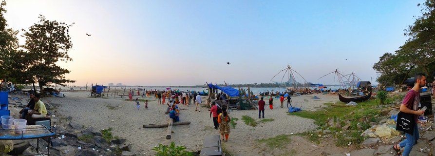 Wide beach scene showing multiple large Chinese fishing nets on the water at sunset, with people walking on sand, boats pulled ashore, and trees framing the edges of the frame