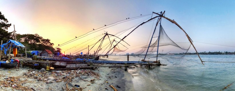 Multiple large Chinese fishing net structures along waterfront at sunset with warm orange sky, calm water, and shoreline with small buildings in Fort Kochi, India