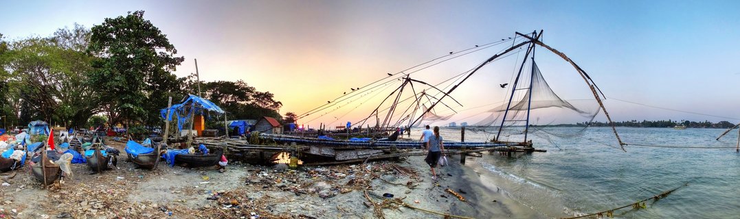Large wooden Chinese fishing net structures along a waterfront at sunset with people walking on shore, fishing boats with blue tarps, and warm orange sky over water in Kochi, India