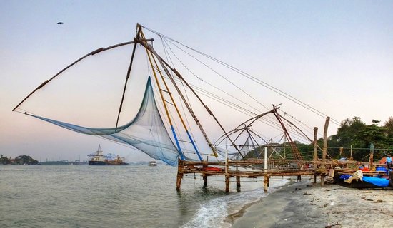 Large wooden Chinese fishing nets suspended over water at Kochi harbor with cargo ship in background and sandy beach in foreground
