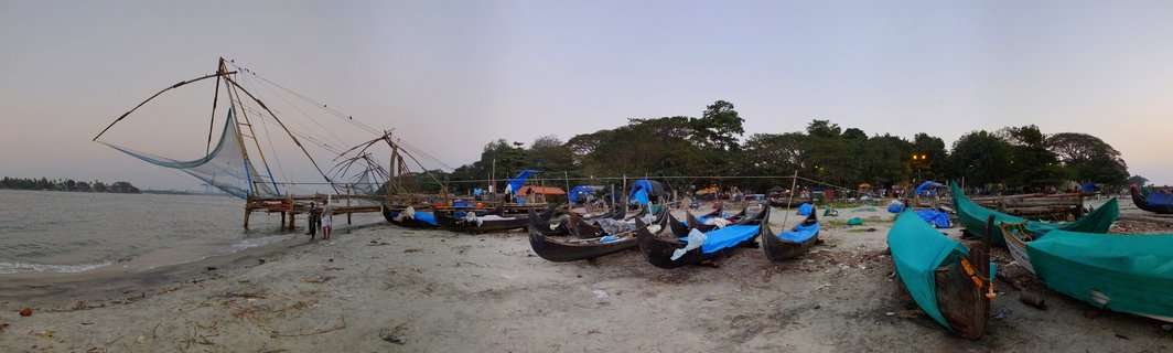 Large Chinese fishing nets on wooden frames along a beach with multiple small fishing boats covered in blue and green tarps on sand, trees in background, twilight sky