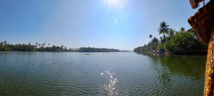 Wide waterway in Kerala backwaters with palm trees on both shores, clear blue sky, sun reflecting on water surface, thatched houseboat roof visible on right edge