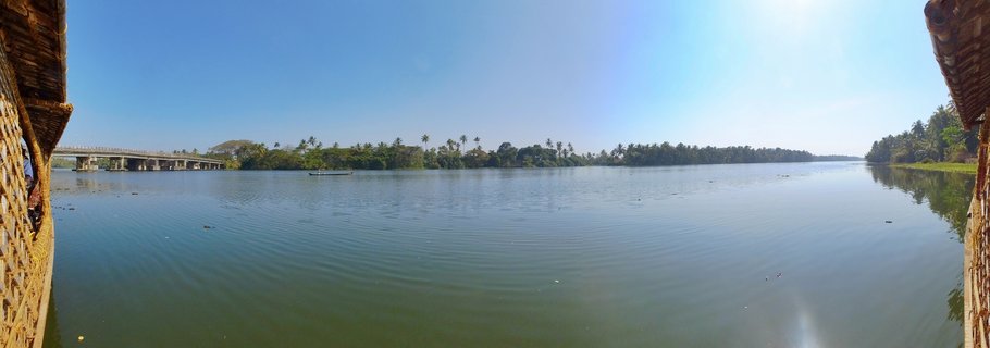 Wide waterway scene showing calm backwaters with a concrete bridge on the left, coconut palm trees along the shoreline, and woven bamboo houseboat structures visible at the frame edges under a clear blue sky