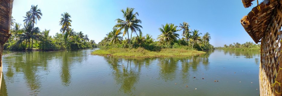 Wide panoramic photograph of Kerala backwaters showing calm green water with coconut palm trees along both banks, a small grassy island in the center, and portions of a woven houseboat visible at the frame edges under a clear blue sky