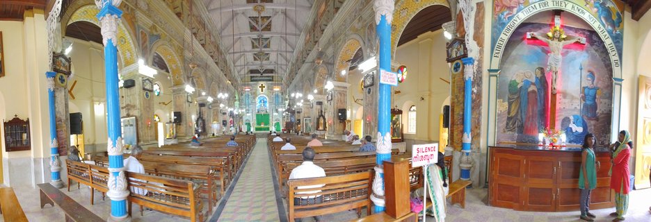 Interior view of a Catholic church showing rows of wooden pews, blue and white decorative columns, religious murals on walls, and people sitting and standing inside the nave