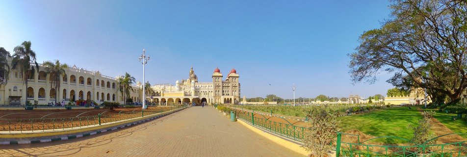 Wide view of Mysore Palace showing the ornate Indo-Saracenic building with red domes, cream-colored facades with arched colonnades, green lawns, paved pathway, and clear blue sky