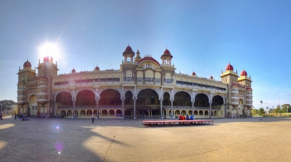 Wide view of Mysore Palace showing the ornate Indo-Saracenic building with red domes, arched colonnades, and cream-colored stone facade against a clear blue sky with visitors in the foreground courtyard