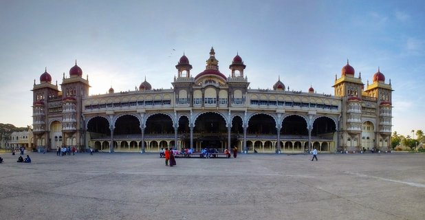 Wide view of Mysore Palace showing cream-colored building with red domes, multiple arched colonnades, and people walking in the paved courtyard