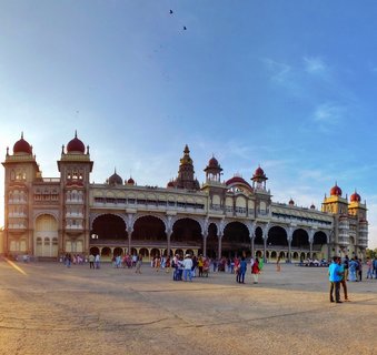 Wide view of Mysore Palace showing the ornate Indo-Saracenic building with red domes, arched colonnades, and cream-colored stone facade with tourists walking in the paved courtyard under a clear blue sky