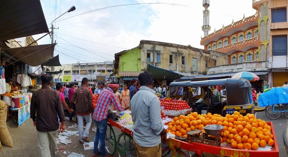 Busy street market in India with fruit vendors selling oranges from red carts, shoppers walking between stalls, colorful buildings and mosque minaret in background