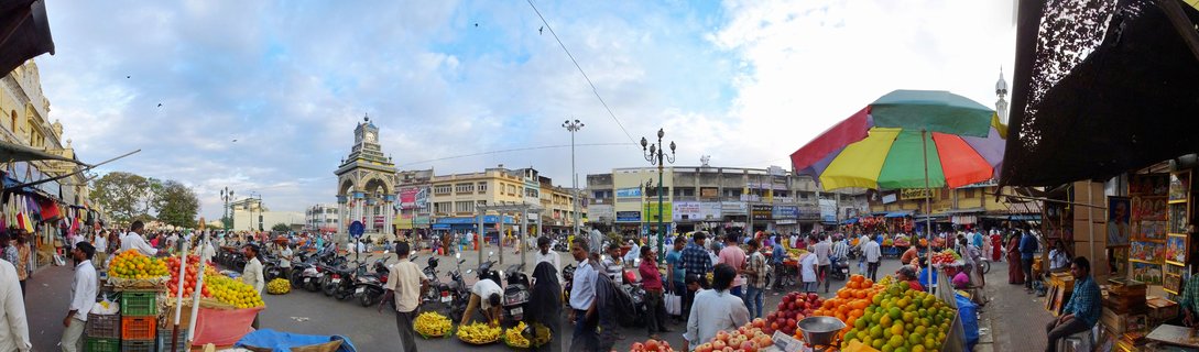 Busy street market in Mysore India with colorful fruit vendor stalls, rainbow umbrella, clock tower arch monument, crowds of people shopping, motorcycles parked, and multi-story buildings under blue sky with clouds