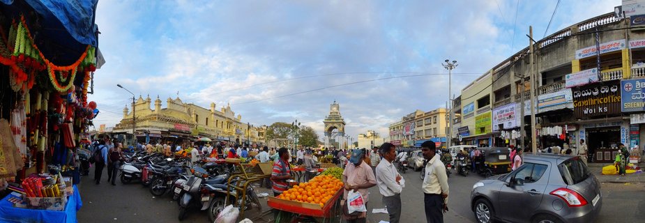 Busy street market in Mysore, India with fruit vendors, flower garlands, motorcycles, and Mysore Palace visible in the background