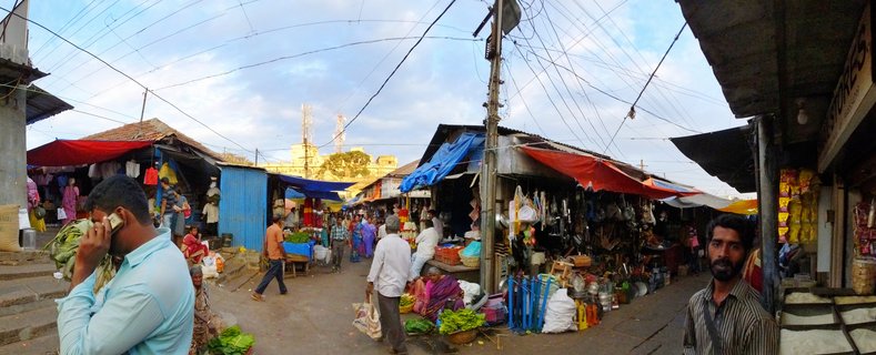 Busy outdoor street market in India with vendor stalls under red and blue tarps, people shopping and walking, vegetables and goods displayed, overhead electrical wires visible against cloudy sky