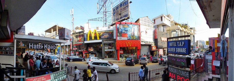 Wide street view of a busy commercial area in Bangalore showing multiple storefronts including McDonald's restaurant, coffee shop, hotel, pedestrians walking, cars and motorbikes on the road, billboards and signage, clear blue sky above