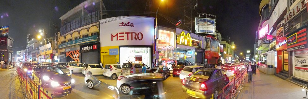 Busy night street scene in Bangalore showing heavy car traffic, illuminated Metro supermarket and McDonald's restaurant, pedestrians on sidewalks, and multiple commercial billboards under street lighting