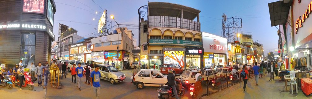 Wide panoramic view of a busy Bangalore street at dusk showing pedestrians walking, cars and scooters in traffic, and multi-story buildings with illuminated commercial signs including McDonald's and banks