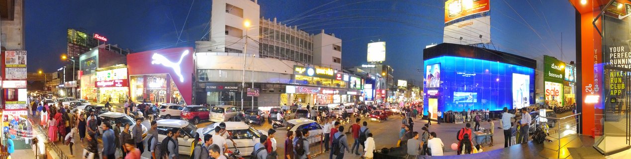 Wide panoramic view of a crowded city street in Bangalore at dusk with illuminated Puma store, bright blue LED billboard, multiple retail shops, parked cars, and numerous pedestrians walking along the commercial district