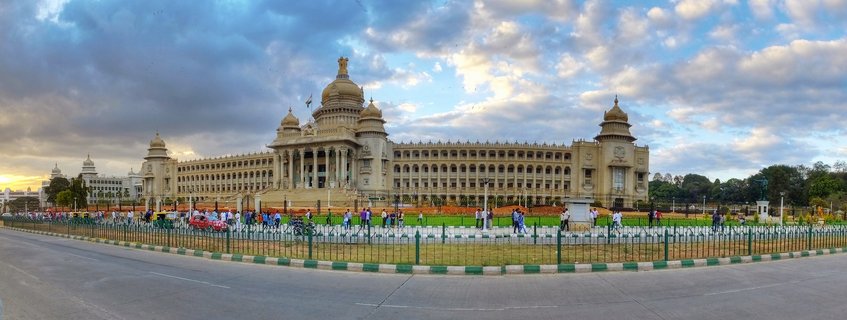 Wide panoramic view of Vidhana Soudha, the Karnataka State Legislature building in Bangalore, India, showing the ornate Indo-Saracenic granite facade with central dome and carved pillars, illuminated by warm golden hour sunset light beneath a dramatic sky with blue patches and white-grey cumulus clouds, with manicured green lawn and paved road in the foreground where scattered pedestrians walk.