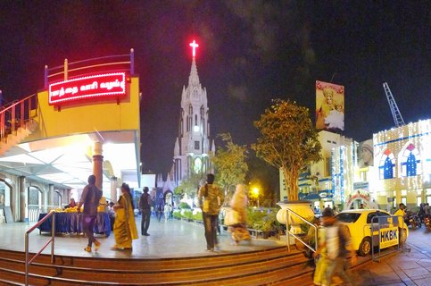 Night photograph of an illuminated Gothic church with a red cross on top, surrounded by lit buildings, pedestrians walking on stairs and plaza, and a yellow taxi in Bangalore, India