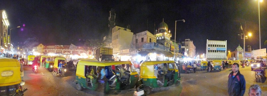 Night street scene in Bangalore showing multiple yellow and green auto-rickshaws in traffic, illuminated shops and buildings, pedestrians, and a mosque with green dome in the background