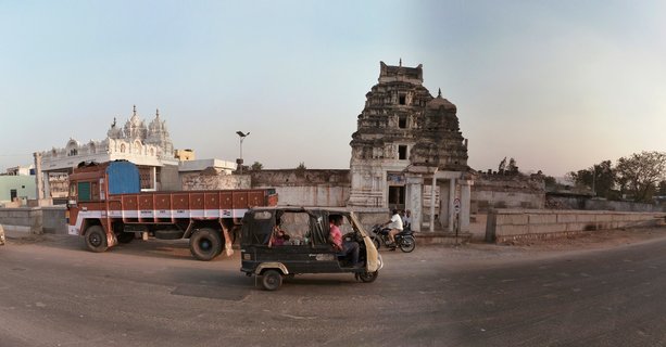 Street scene showing a black auto-rickshaw driving on a road with an ancient stone temple tower and white temple building in the background, cargo truck parked on the left side, Hampi, India