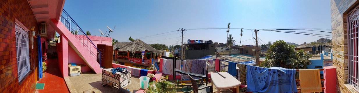 Panoramic rooftop view showing colorful pink and red buildings, laundry hanging on clotheslines, outdoor furniture, satellite dishes, power lines, and palm trees under a clear blue sky in Hampi, India