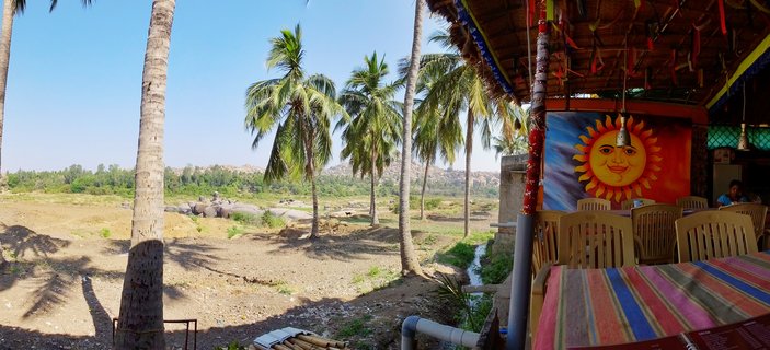 Outdoor cafe with colorful sun mural, plastic chairs and striped tablecloths under palm trees, overlooking dry rocky riverbed with boulders and hills in Hampi, India